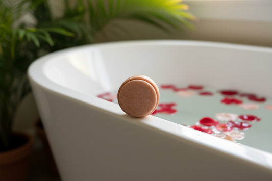 Pink circular shampoo bar on a white tub, and rose petals in the water.
